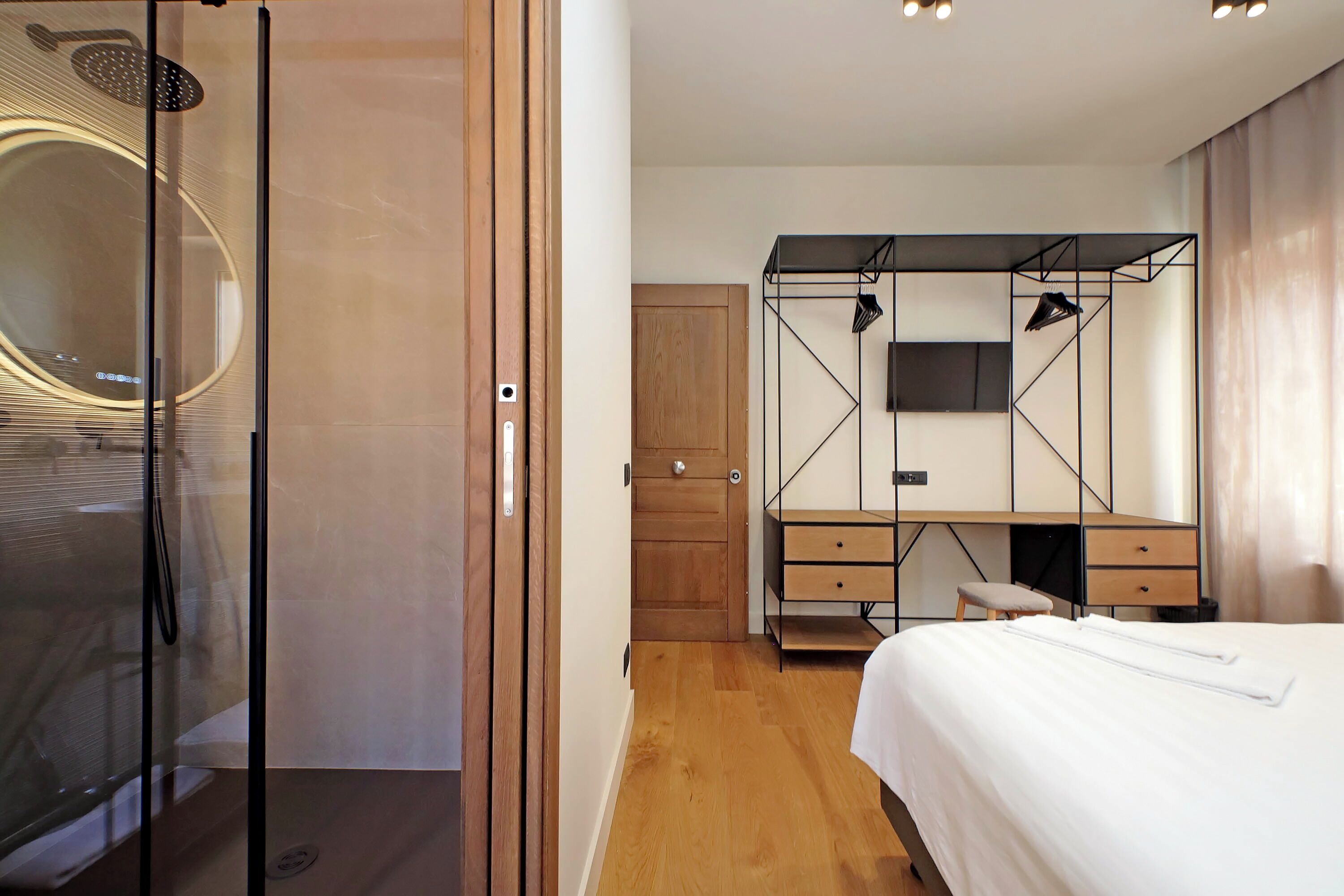 A modern bedroom with a glass shower enclosure, a wooden wardrobe, a bed with white bedding, and a mounted TV.