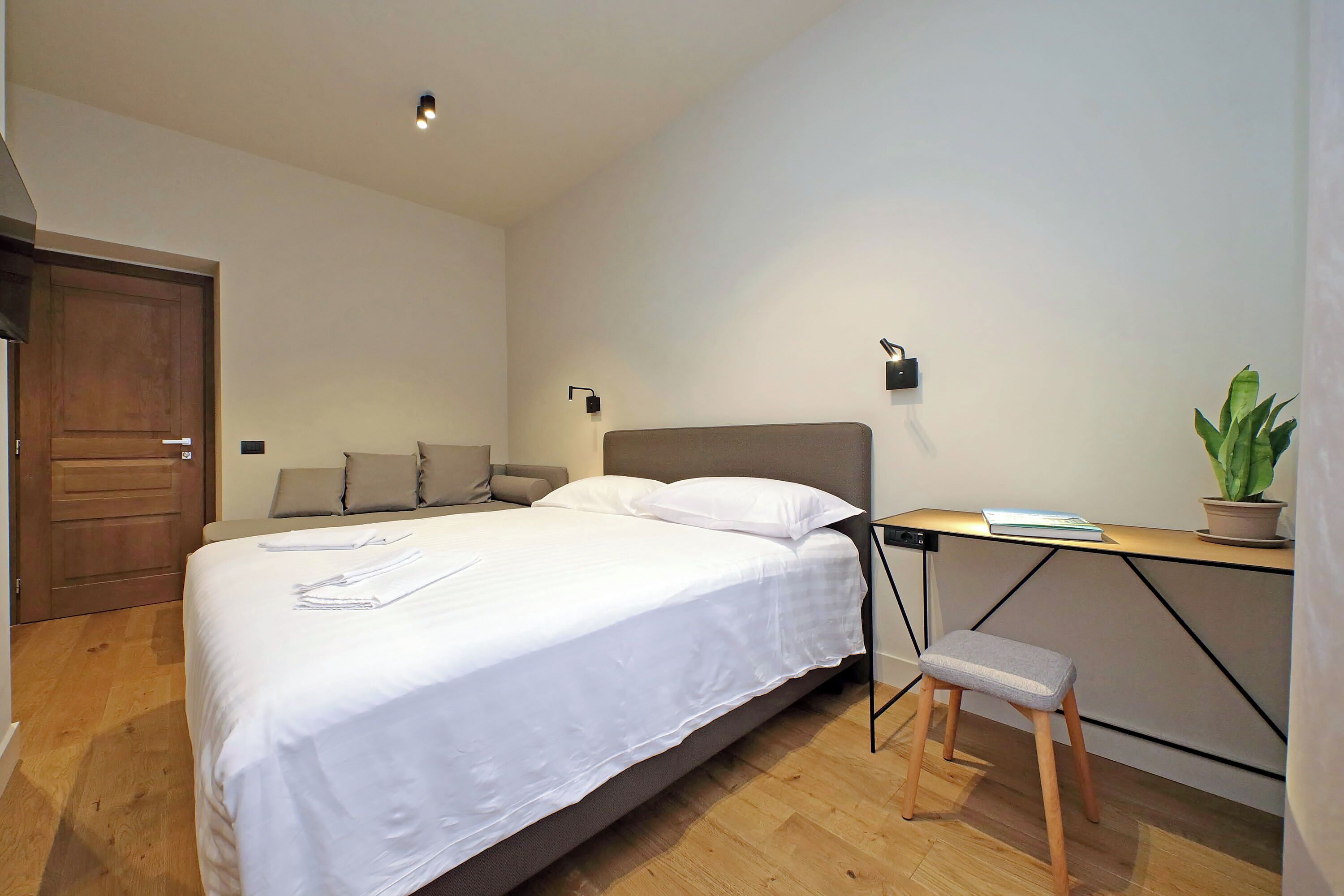 A bedroom with a bed, a desk, a chair, a potted plant, and a wooden door.