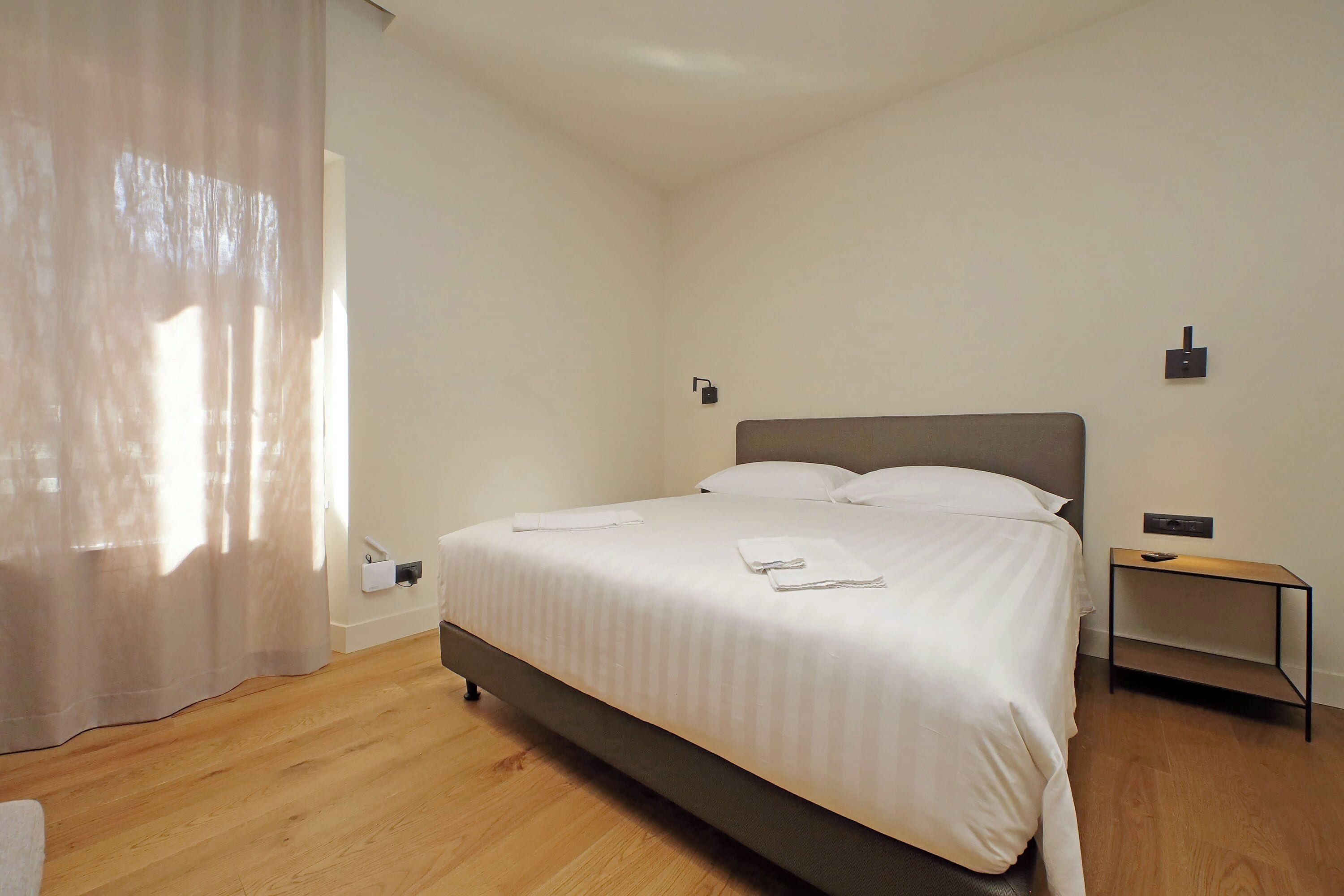 A neatly made bed with white linens, a wooden floor, a bedside table with a lamp, and a wall-mounted switch.