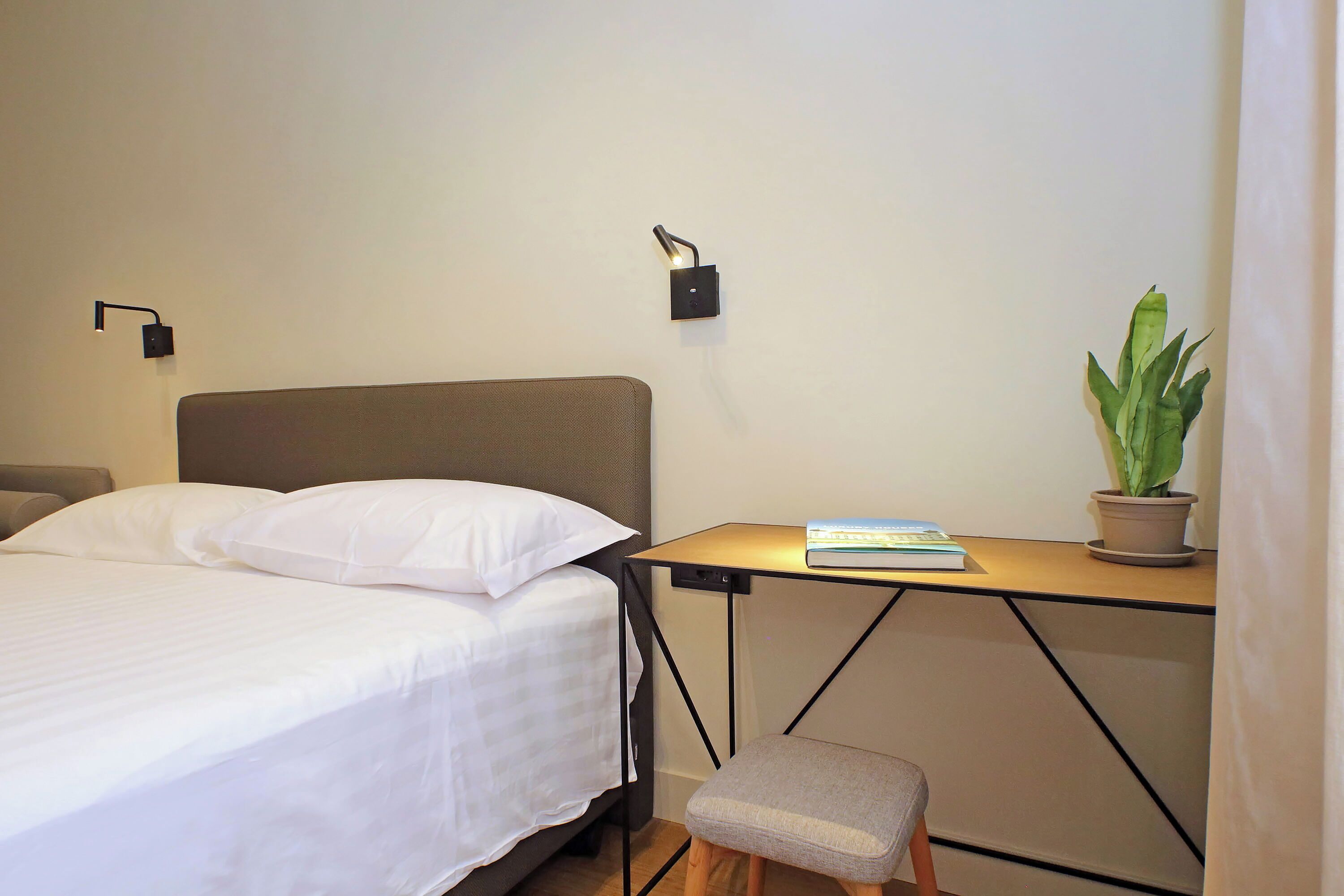 Un lit bien fait, avec du linge de lit blanc, un petit tabouret en bois, une table avec un livre, une plante en pot et deux lampes fixées au mur.
