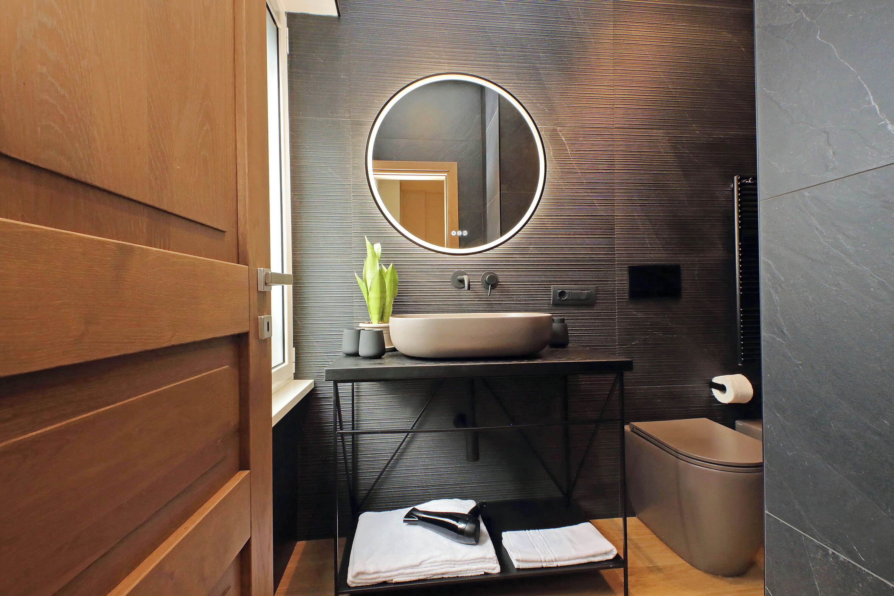 Een moderne badkamer met een houten wastafelmeubel, een ronde spiegel, een wastafel, toilet en een prullenbak.