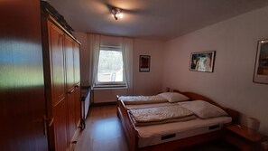 Makuuhuoneessa on puinen vaatekaappi, ikkunassa verhot, kaksi sänkyä tyynyineen sekä kaksi kehystettyä kuvaa seinällä.