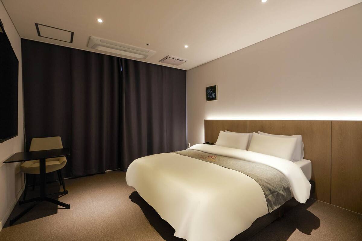 Quarto de hotel com uma cama grande, uma mesa com cadeira e cortina escura.