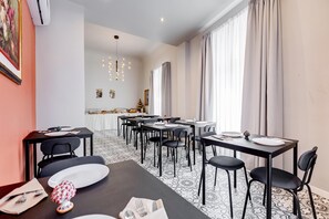 A dining area with tables set for four, chairs, a chandelier, and a view of the kitchen in the background.