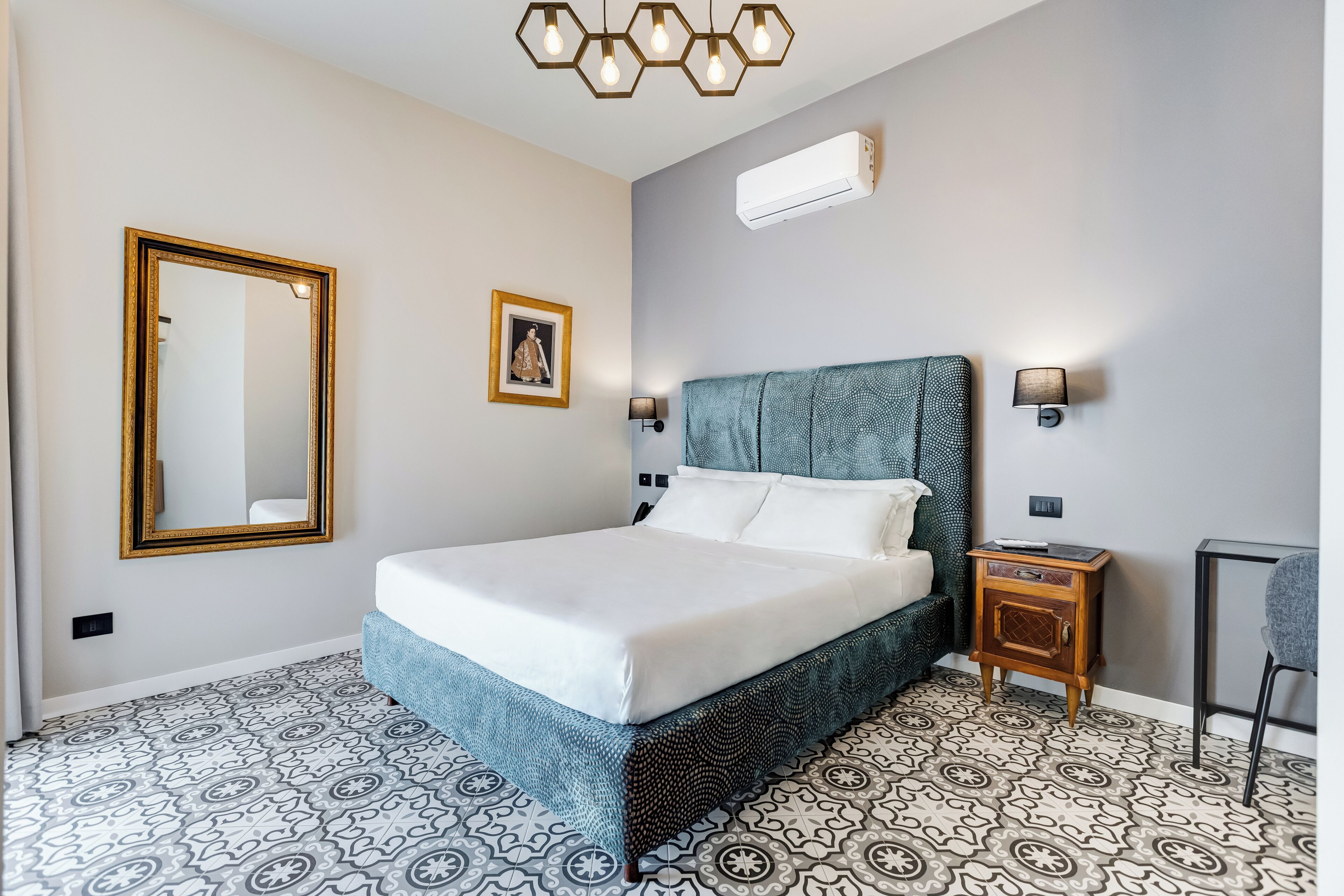 A hotel room with a bed, a nightstand, a wall-mounted air conditioner, a mirror, and patterned flooring.