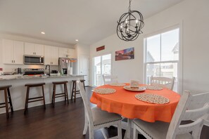 Cocina luminosa con armarios blancos, un microondas y un refrigerador. Hay una zona de comedor con una mesa dispuesta para cuatro, sillas blancas y un mantel naranja. Un candelabro cuelga sobre la mesa.