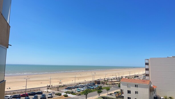 Blick auf einen Strand von einem Gebäude aus, im Vordergrund ein Parkplatz und eine Straße.
