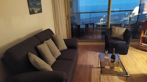 Ein Wohnzimmer mit einem dunkelgrauen Sofa, einem Glastisch mit einer Flasche und Gläsern sowie Meerblick durch große Fenster.