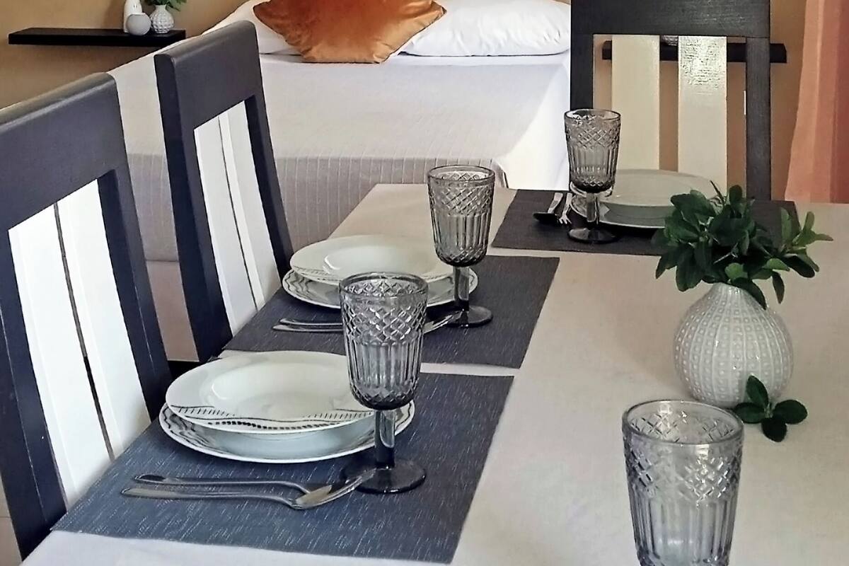 A dining area with a table set for two, including plates, glasses, and cutlery. There is a ceiling fan above, and a bed with a pillow in the background.
