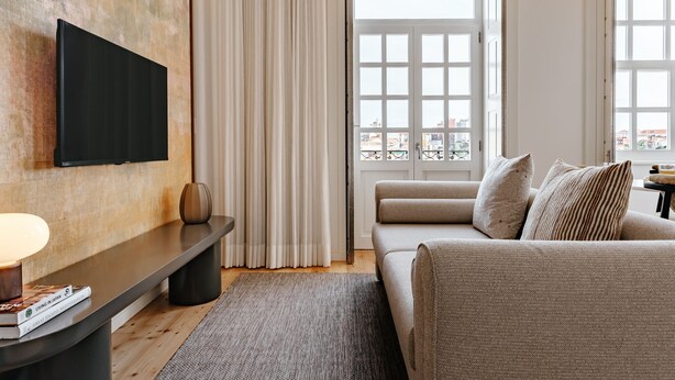 Een moderne woonkamer met een flatscreen televisie aan de muur, een bank met kussens, een bijzettafeltje met een lamp en boeken, en een groot raam met gordijnen.