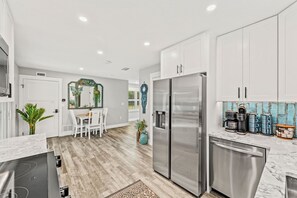A modern kitchen with white cabinets, a stainless steel refrigerator, and a dining area with a table and chairs. - Shell Haven | Luxury Beachfront Villa | Heated Pool + Spa | Steps From The Sand (Melbourne Beach)