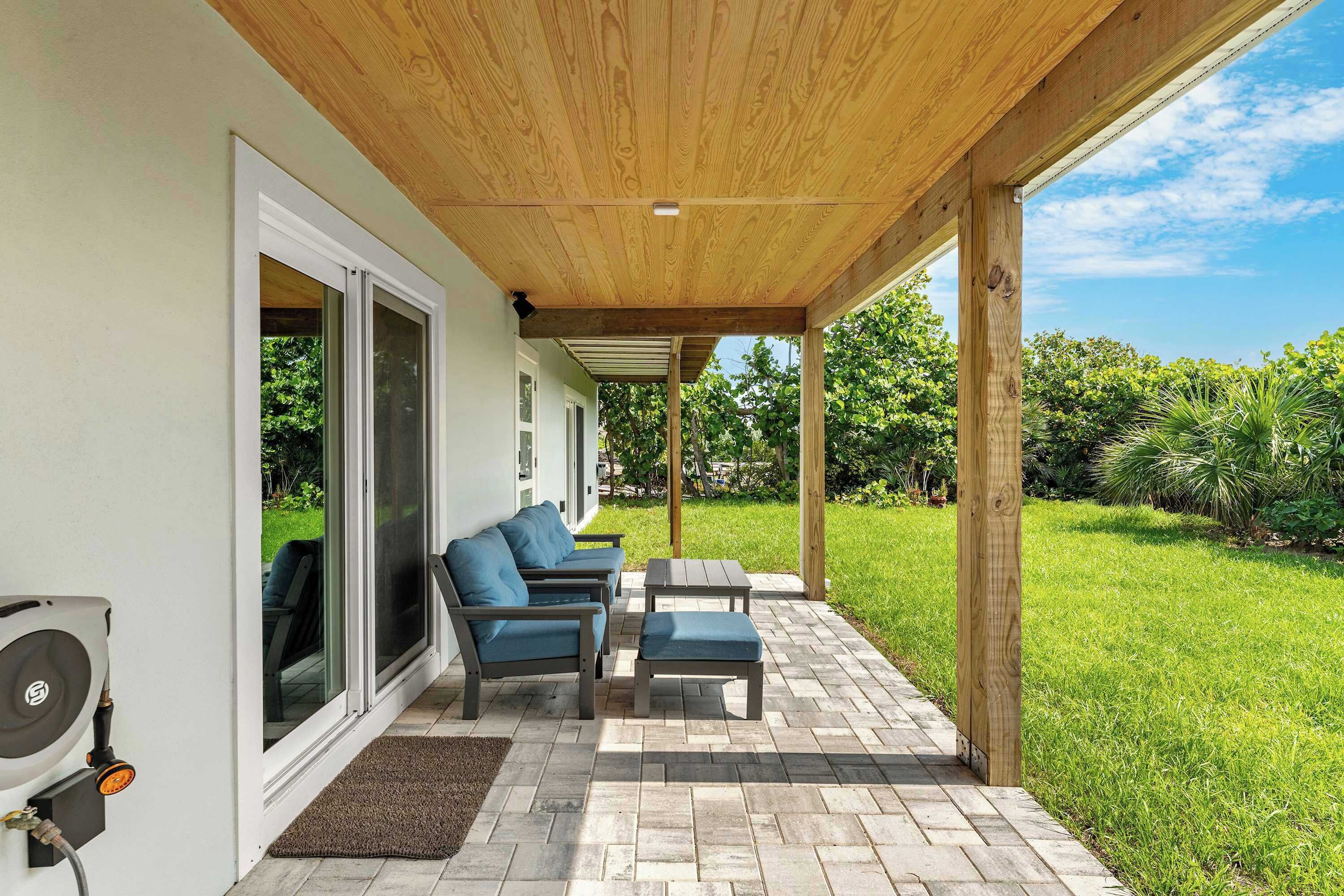 An outdoor patio with a wooden ceiling, a seating area with a sofa and chairs, and a view of a green lawn.