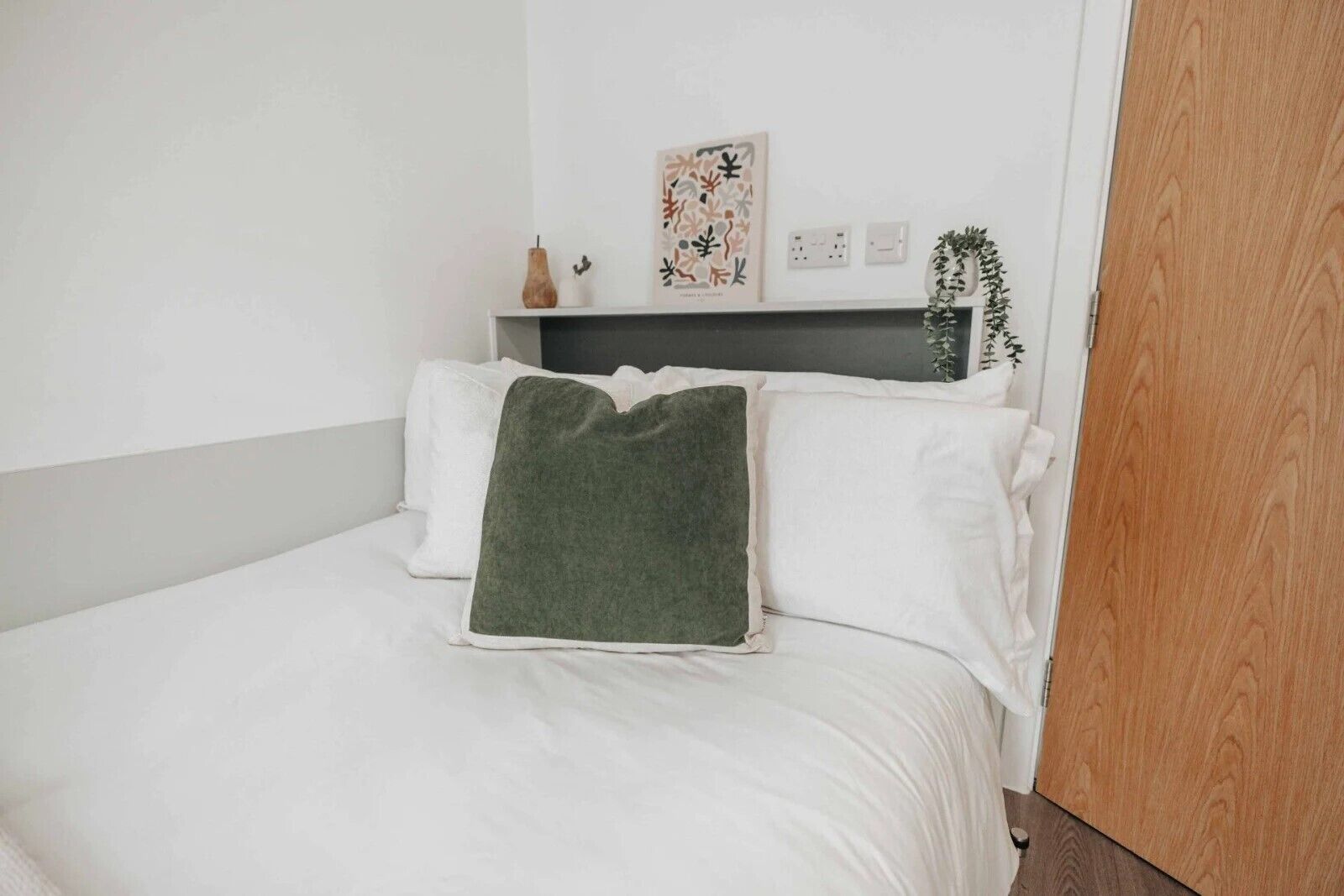 A bed with white bedding and a green throw pillow, a small shelf with decorative items, and a wall-mounted light switch.