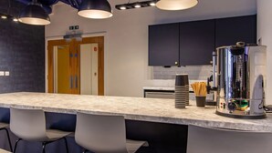 A modern kitchen with a bar area, featuring a large coffee machine, a blender, and a kettle. There are three bar stools and a counter with a marble top. - Alma Court - Campus Accommodation (Canterbury)