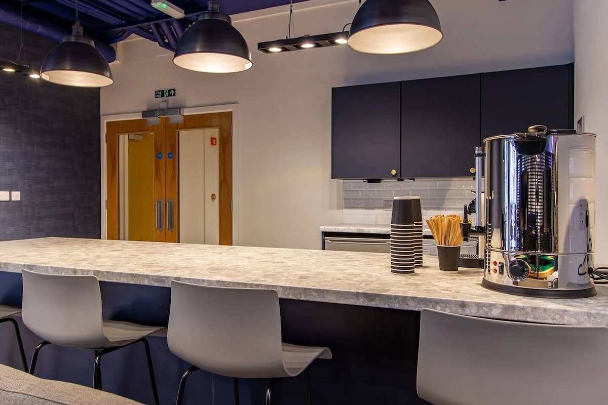 A modern kitchen with a bar area, featuring a large coffee machine, a blender, and a kettle. There are three bar stools and a counter with a marble top.