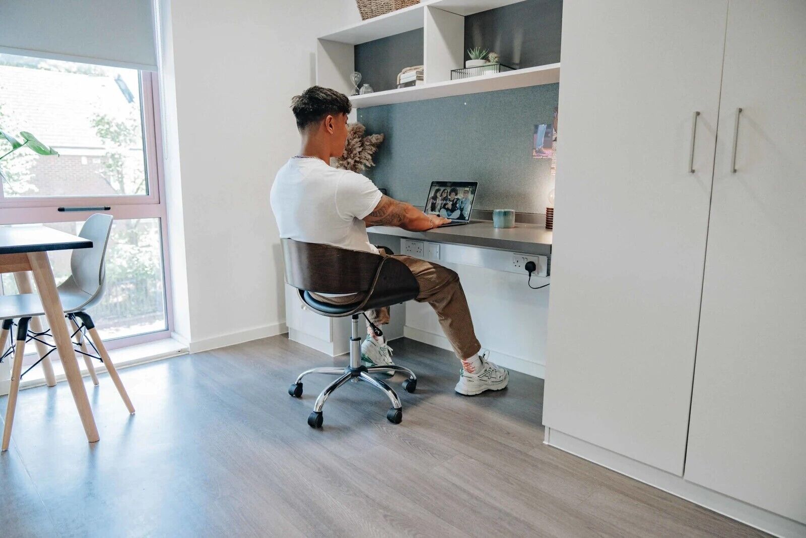 A person sitting at a desk in a room with a window, a chair, and a cabinet.