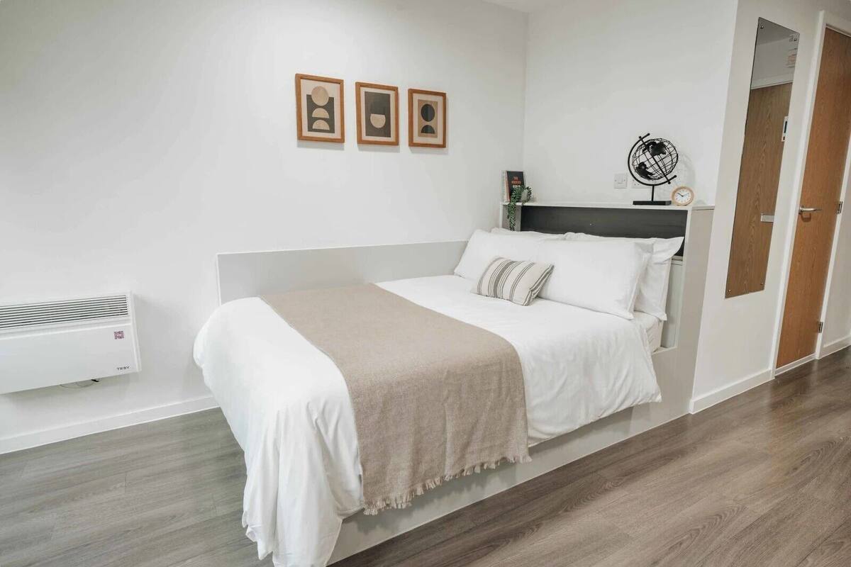 A neatly arranged bedroom with a bed, a bedside table, a wall-mounted heater, and three framed pictures on the wall.