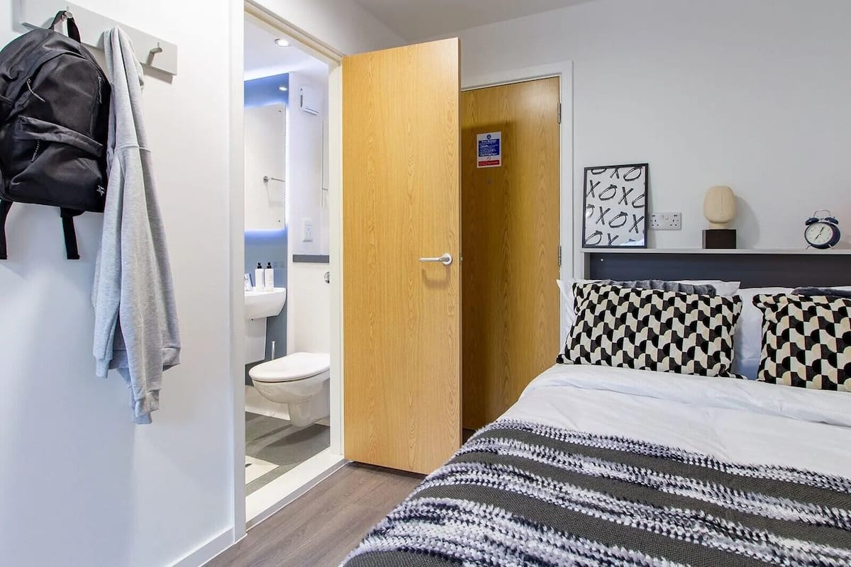 A bedroom with a bed, checkered pillows, a wooden door, and a bathroom visible through an open door.