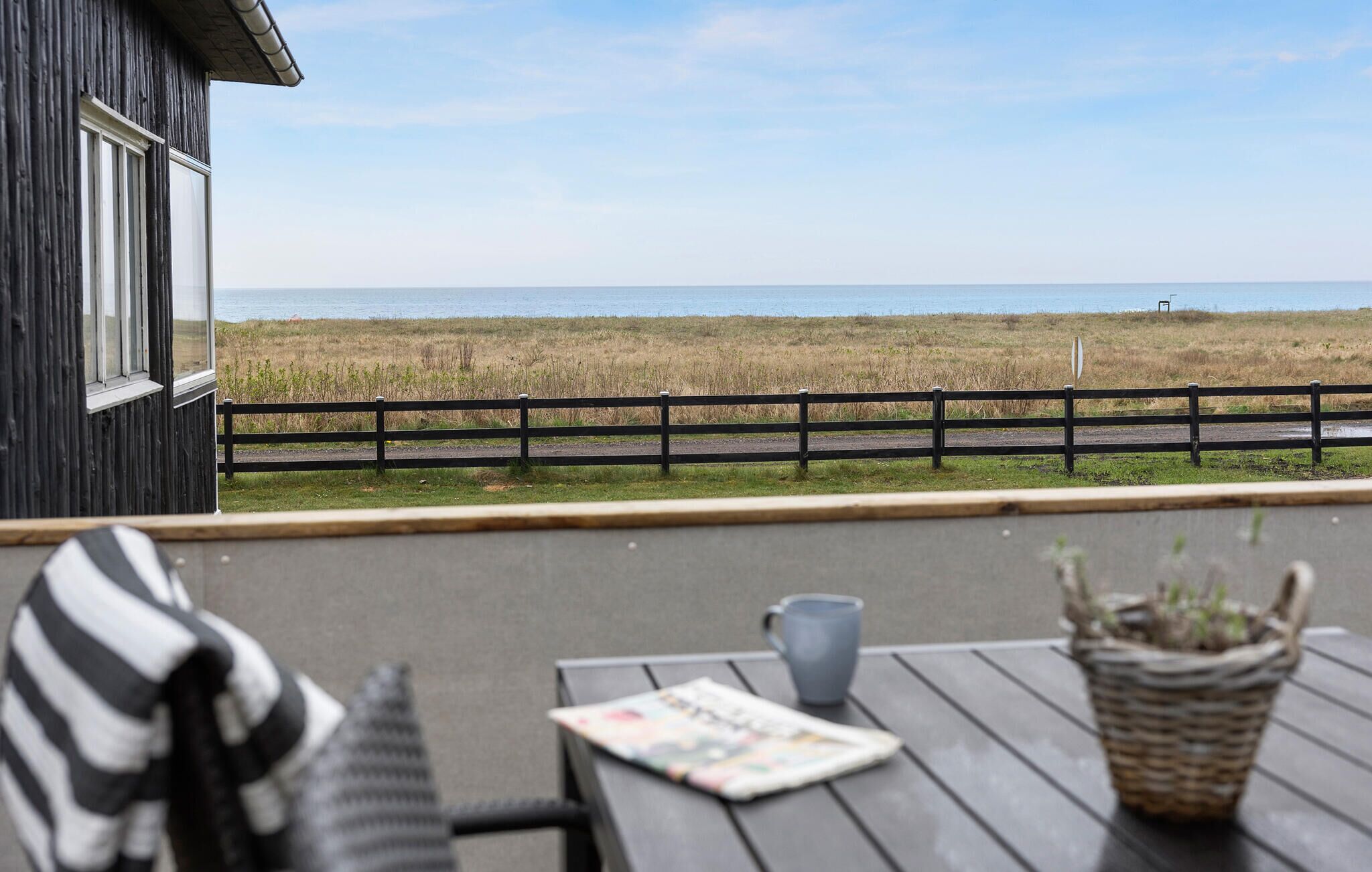 En træterrasse med udsigt til stranden, et hegn, et bord med en avis, en kop og en potteplante.