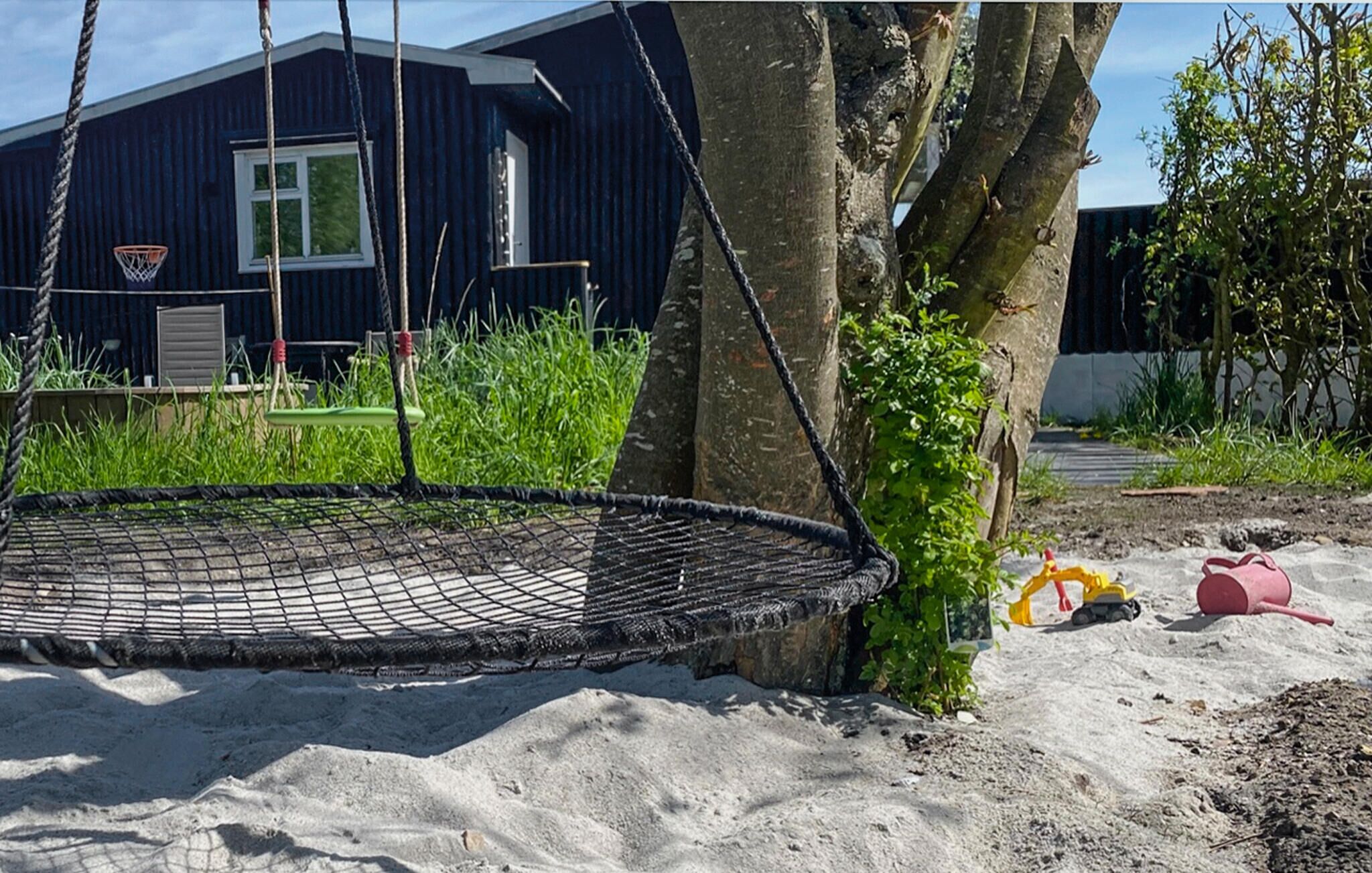 A hammock swing attached to a tree, a sandbox, and a building in the background.