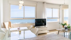 A modern living room with a large TV, glass dining table, and seating. It features large windows with ocean views. - Beach front apartment (San Luis de Sabinillas)