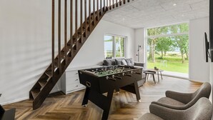 A modern living room with a wooden staircase, a foosball table, and a view of a green landscape through large windows.
