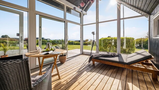 A sunlit outdoor deck with a wooden table, a wicker chair, and a cushioned seating area.
