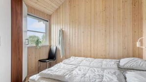 Un dormitorio con paneles de madera, una cama con ropa blanca, una silla negra y una ventana con vistas a zonas verdes.