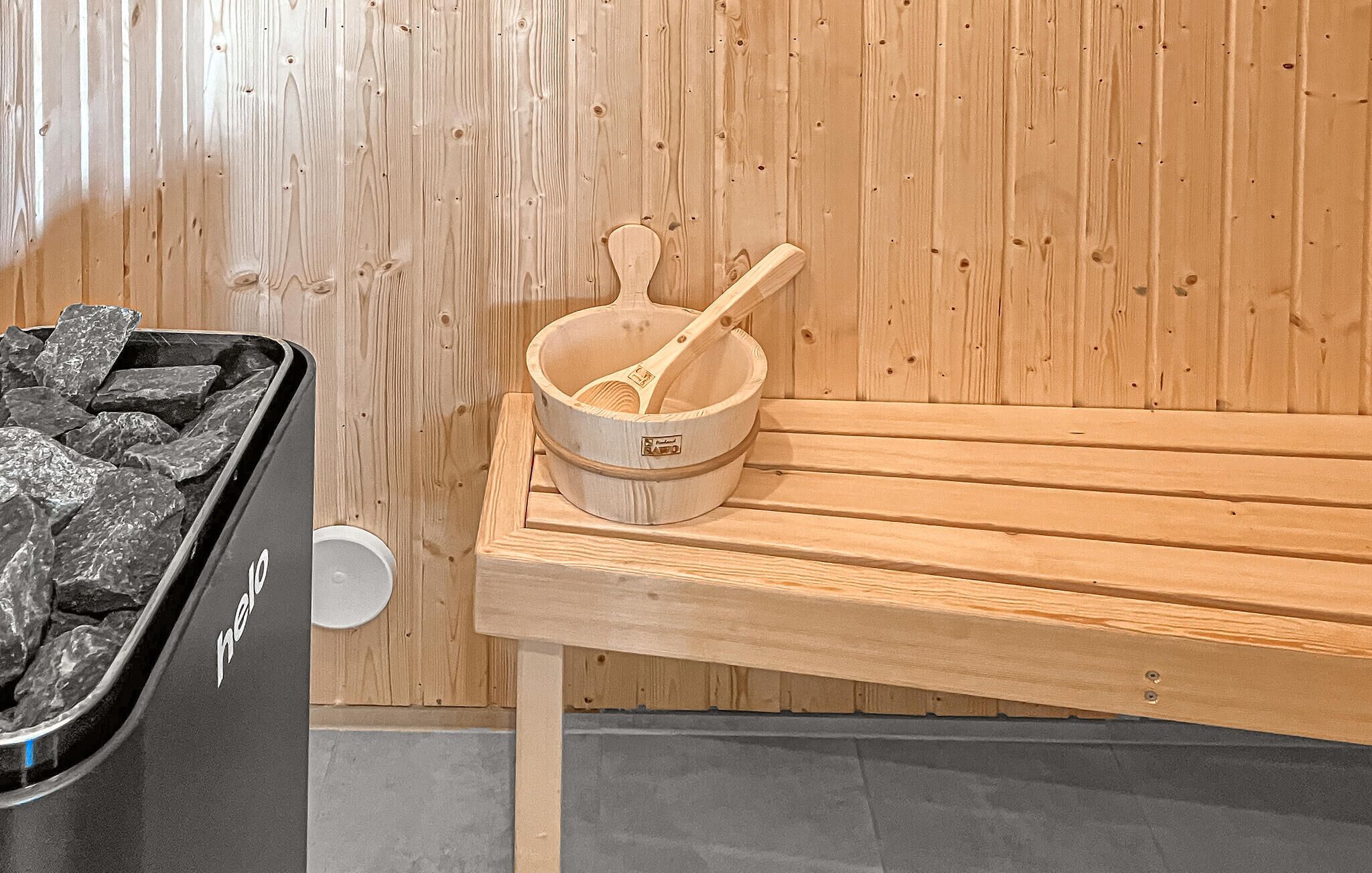 A sauna room with a wooden bench, a wooden bucket with a ladle, and a trash bin labeled "help."
