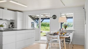 Et moderne køkken med hvide skabe, en indbygget ovn og en kaffemaskine. Der er et spiseområde med et bord dækket til to personer, et ur på væggen og udsigt til grøn vegetation udenfor.