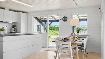 A modern kitchen with white cabinets, a built-in oven, and a coffee machine. There is a dining area with a table set for two, a clock on the wall, and a view of greenery outside.