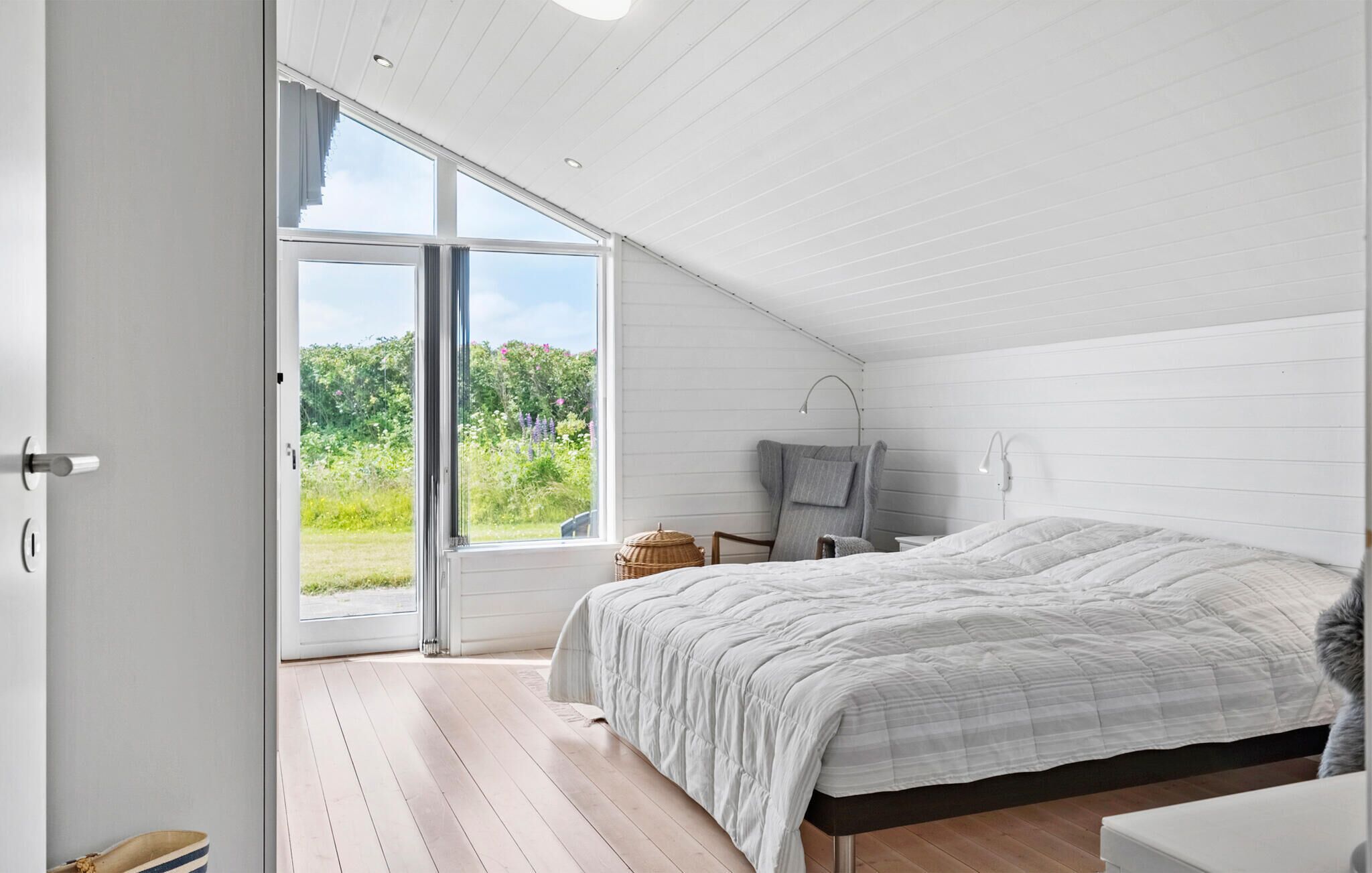 A bedroom with a large bed, a desk, and a chair. There is a window with a view of greenery outside.