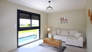 A modern living room with a white sofa, a wooden coffee table, and a view of a swimming pool through a large window. - Lovely home in Puy St Martin (Puy St Martin)
