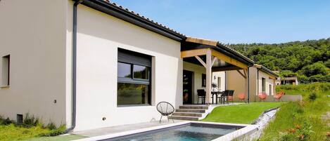 A modern house with a swimming pool, a covered patio, and a lush green landscape.