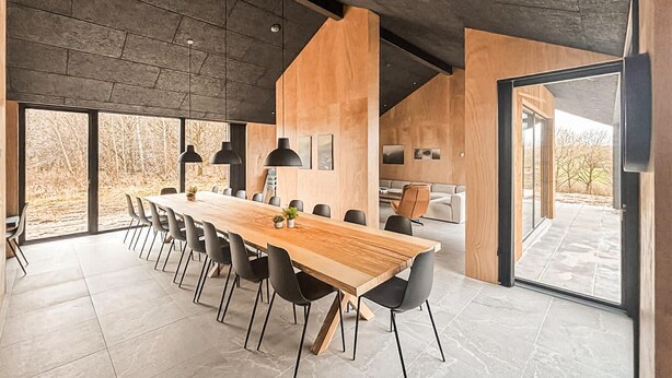 Een moderne eethoek met een lange houten tafel, zwarte stoelen, hanglampen en een uitzicht door grote ramen.