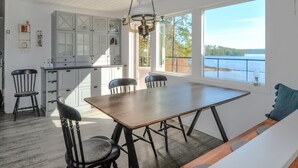 Een eetgedeelte met een houten tafel en vier stoelen, een keuken met witte kasten, en een groot raam met uitzicht op een waterlichaam.