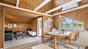 A modern interior with a wooden ceiling, a dining area with a round table and chairs, a sofa, and a bookshelf.