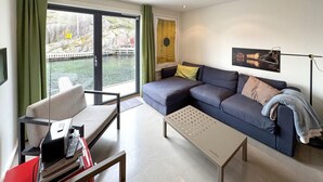 A modern living room with a sofa, a coffee table, and a chair. There is a window with a view of a river and buildings.