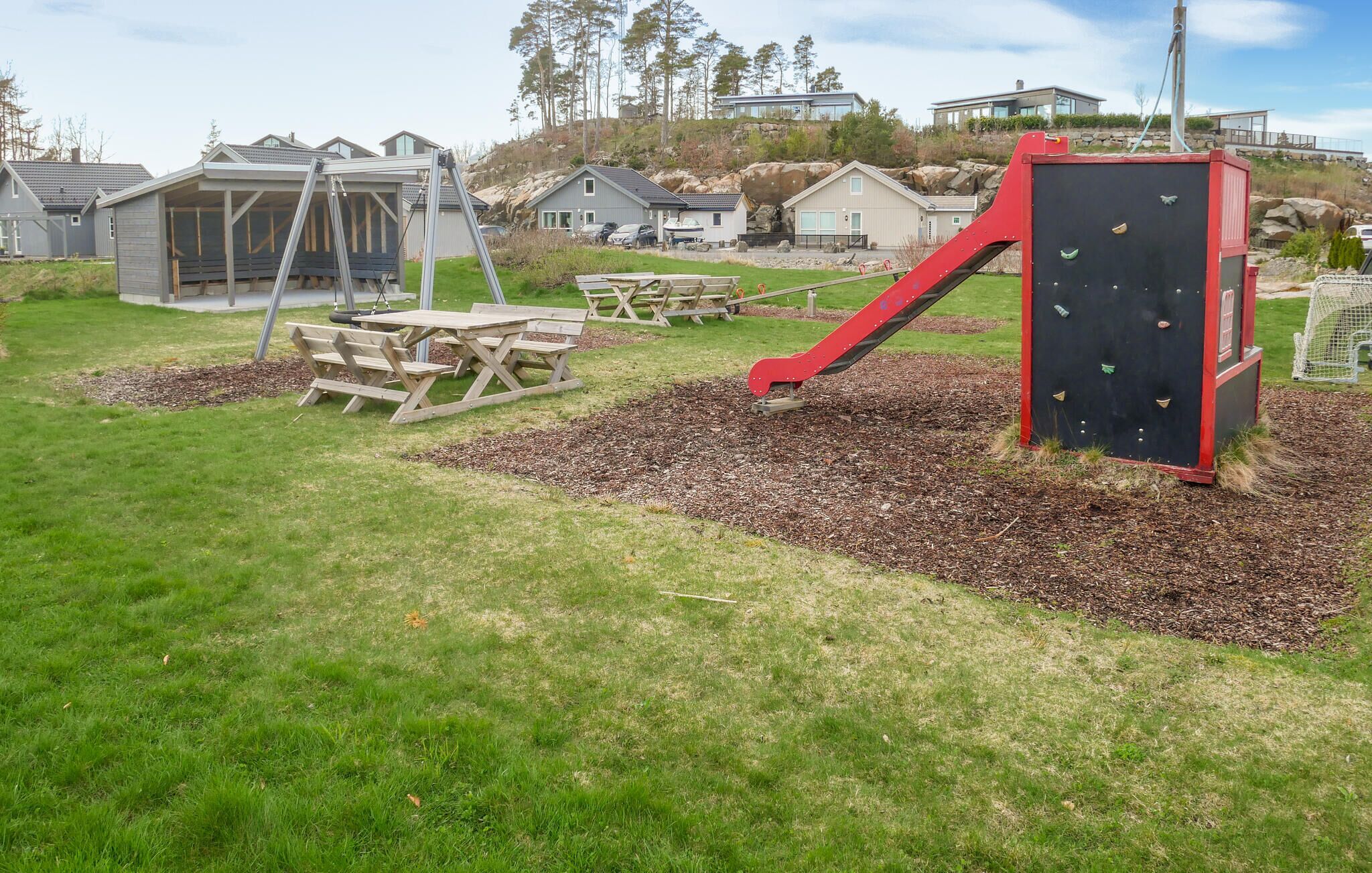 Un parque infantil con pared de escalar roja, mesas de picnic y columpios.
