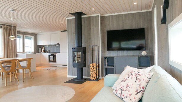 A modern living room with a fireplace, a TV mounted on the wall, and a dining area with a table and chairs. - 4 bedroom cozy home in Færvik (Færvik)
