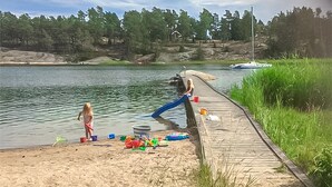 Une jetée en bois s’avance sur un lac dont les rives sont sablonneuses. On aperçoit des bateaux sur l’eau et une maison au loin.