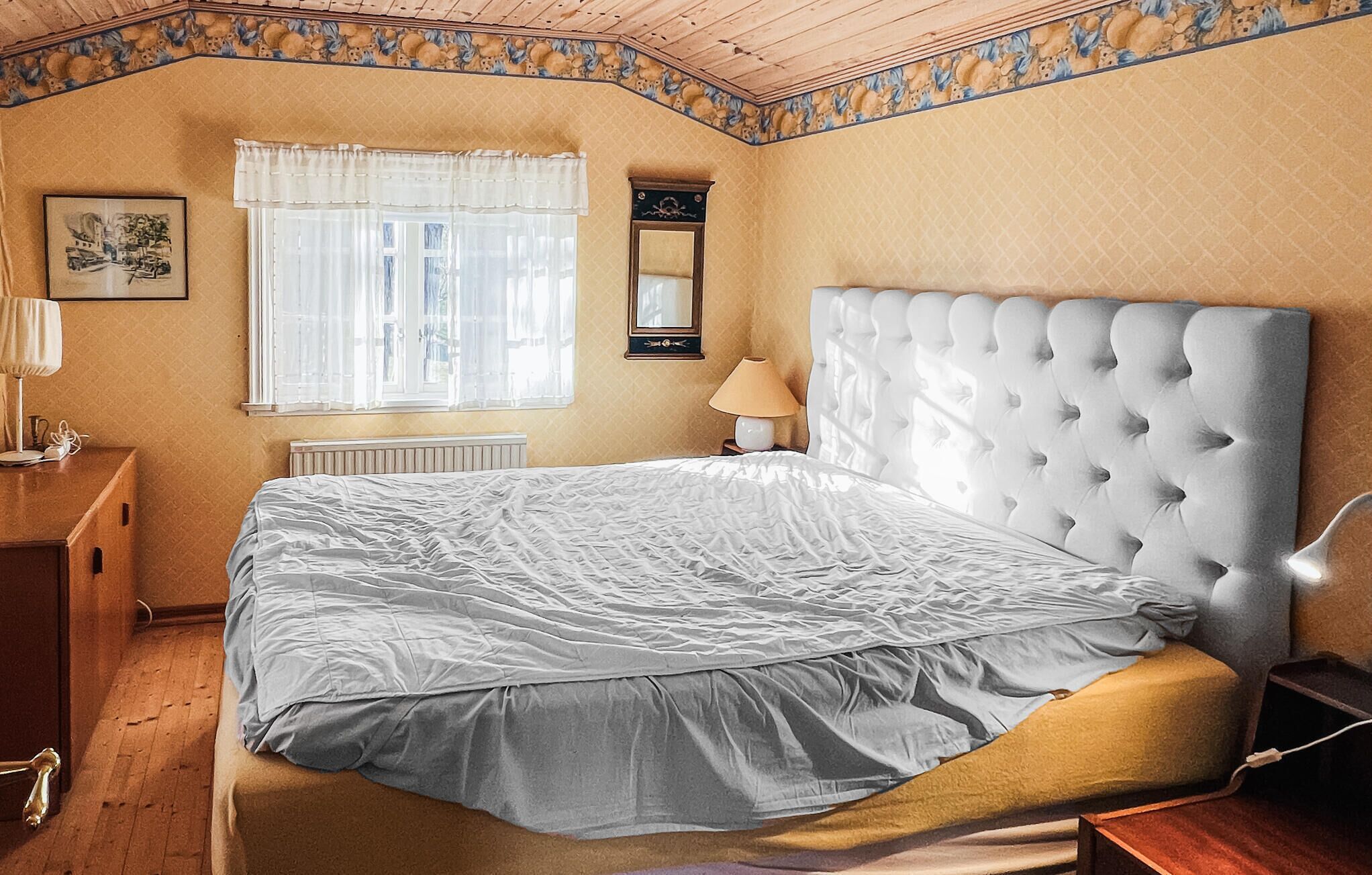 Un dormitorio con una cama grande, una ventana con cortinas translúcidas, una lámpara de noche, un radiador y un cuadro en la pared.