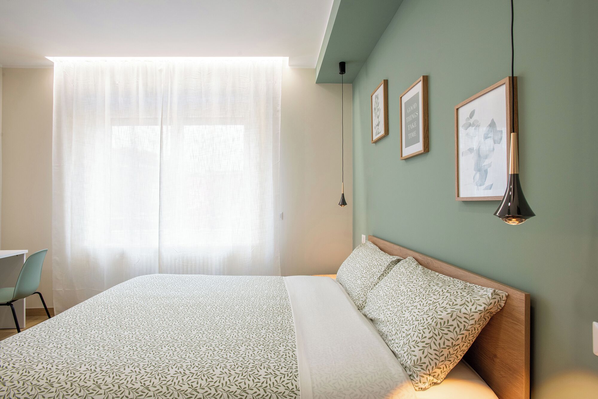 A bedroom with a bed, a chair, a window with sheer curtains, and three framed pictures on the wall.