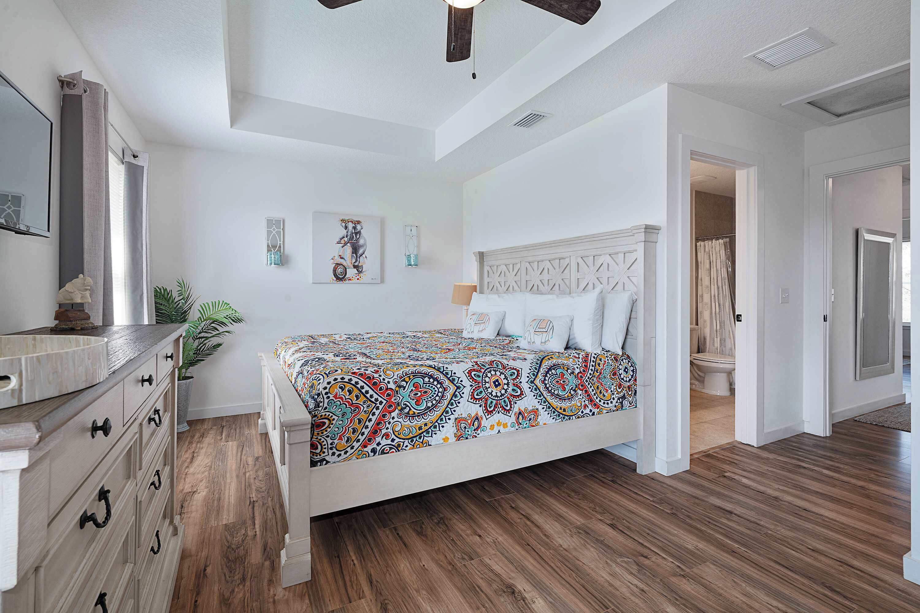 A bedroom with a wooden floor, a bed with a patterned bedspread, a dresser, a mirror, and a ceiling fan.