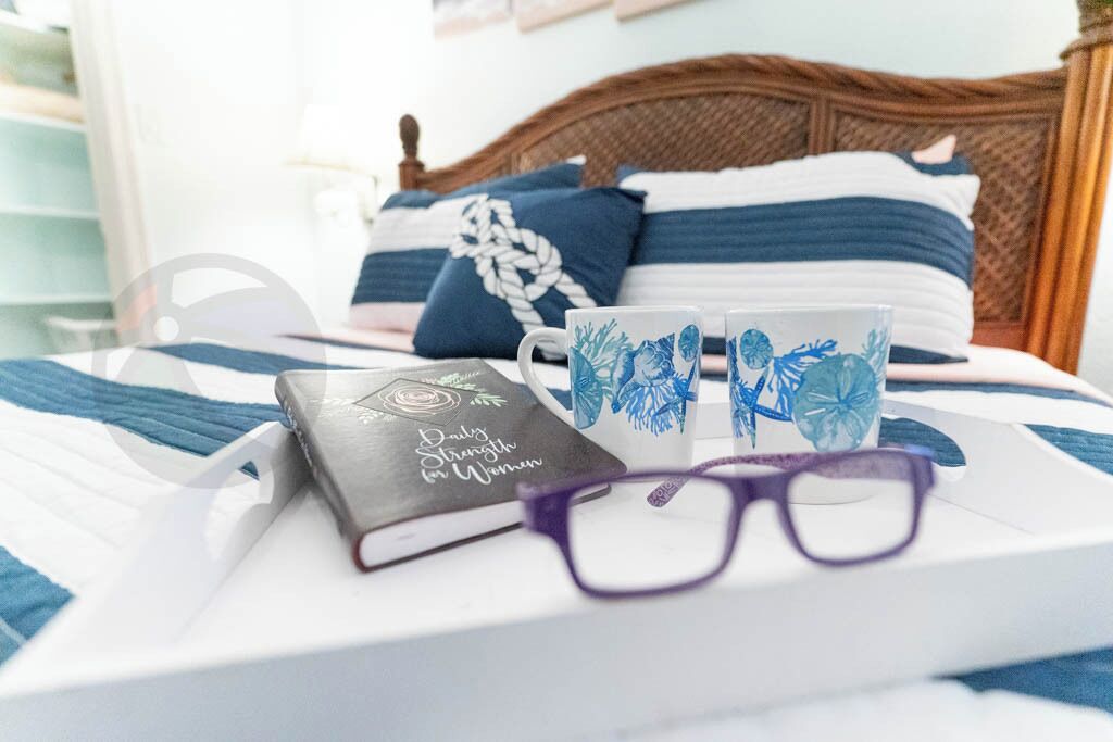 A neatly made bed with blue and white bedding, a book titled "Daily Strength for Women," two decorative mugs, and a pair of glasses on a tray.