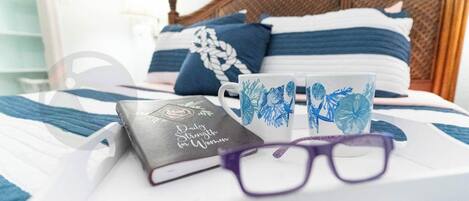 A neatly made bed with blue and white bedding, a book titled "Daily Strength for Women," two decorative mugs, and a pair of glasses on a tray.