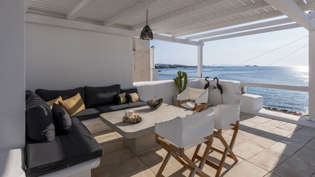 A sunlit outdoor seating area with a sofa, chairs, and a table, overlooking a body of water. - Walkable distance from everything you need (Paros)