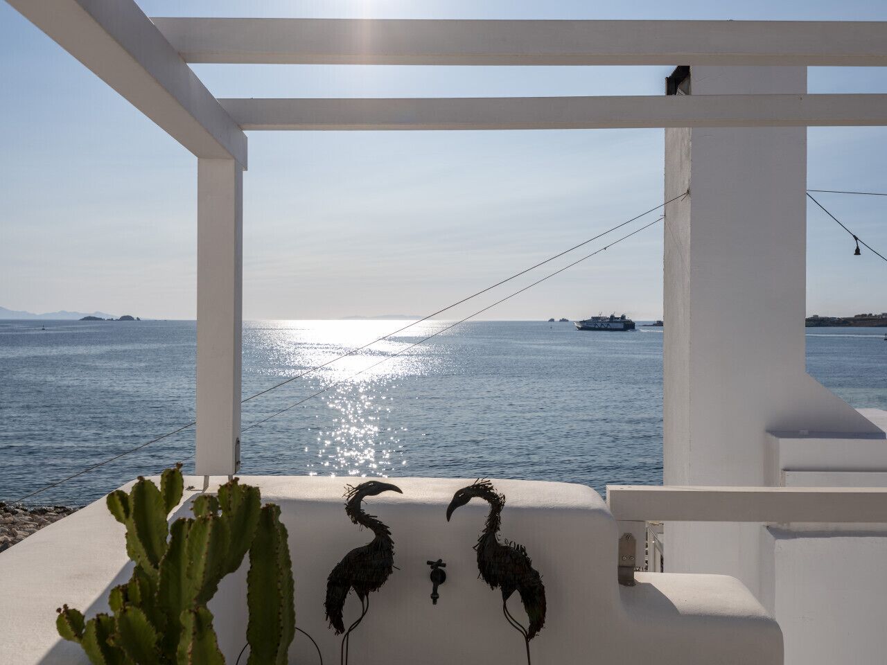 Un balcón blanco con vistas al mar y a islas distantes, con una planta de cactus y dos esculturas de aves.