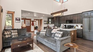 A spacious living room with a grey sofa set, a coffee table, and a small side table. The kitchen features dark cabinets, a stainless steel refrigerator, and a central island with a sink. - 34 Acre Escape~Games~Hiking~Pool Table~Bicycles (Black Hawk)