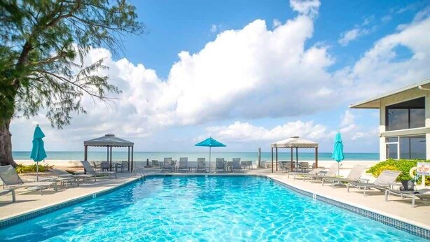 Una piscina con sillas de descanso y sombrillas, vista a la playa y cielo despejado.